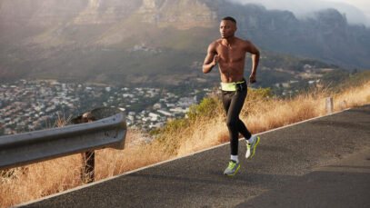 Athletic healthy man runs along road outdoors against mountain background, covers long distance, prepares for marathon. Sporty male exercises down hill, wears sportshoes, leggings, being in good shape carbogel