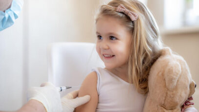 Immunization for children concept. Happy little cute blonde girl holding a toy and getting a flu shot not afraid of the syringe needle. Doctor injecting brave child with Covid-19 vaccine at clinic or hospital poliomielite